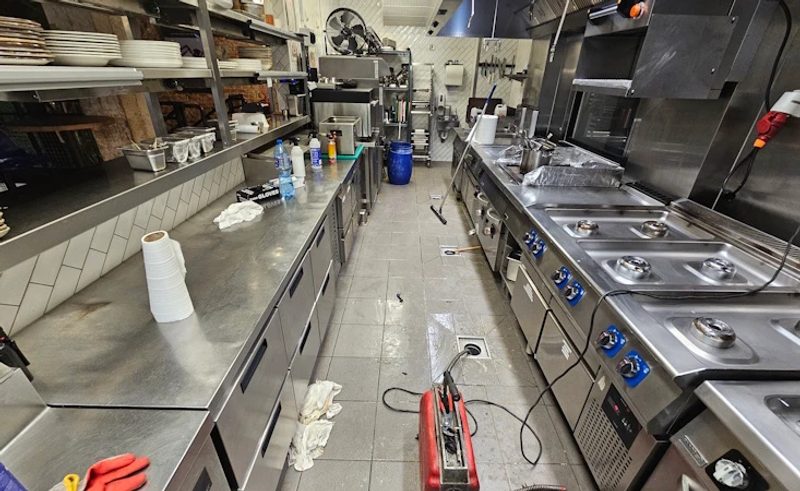 Elektryczna sprężyna do udrażniania kanalizacji w wąskiej kuchni restauracji – spirala wprowadzona do wpustu podłogowego między urządzeniami gastronomicznymi